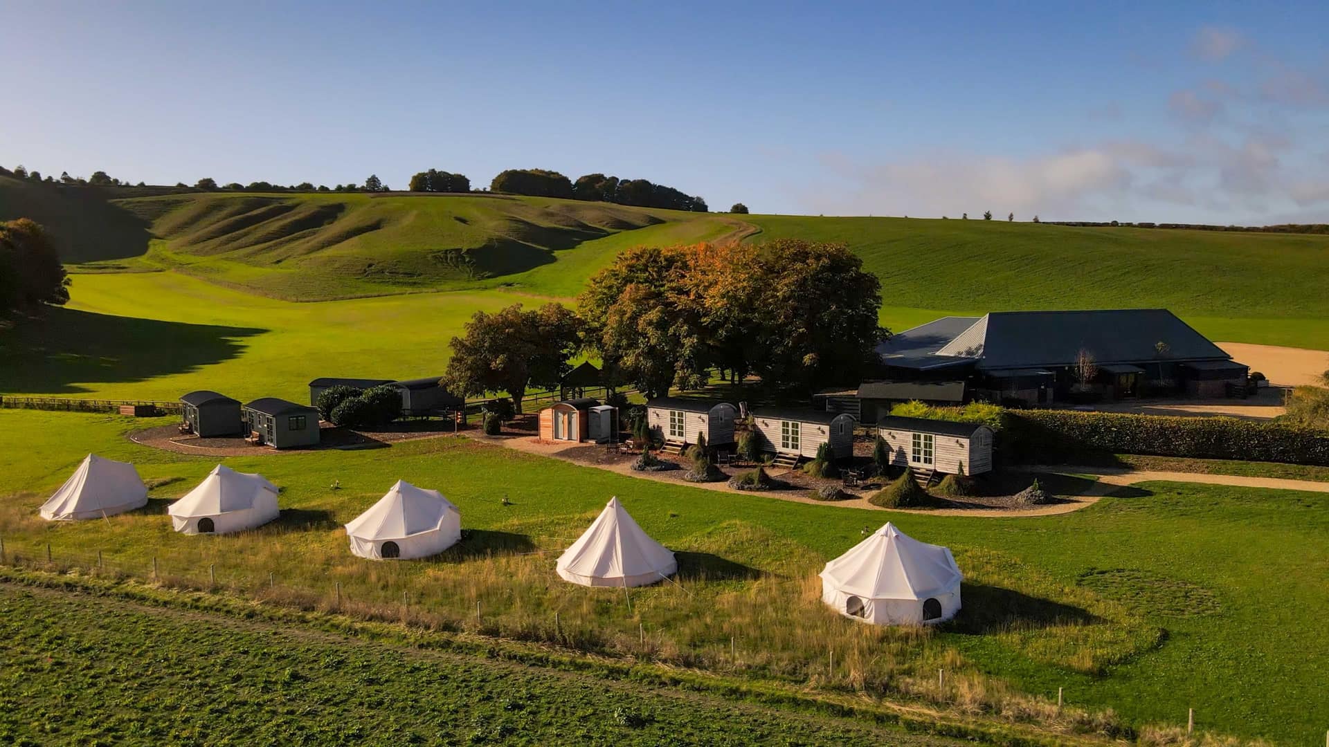 Golden hour aerial over Wellington Barn, the bell tents, shepherd huts and the empty Downs behind