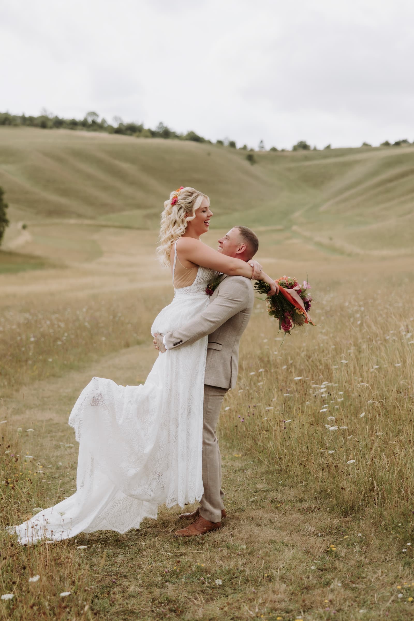 A groom lifting his bride mid spin in the wild flower meadow, the Downs behind